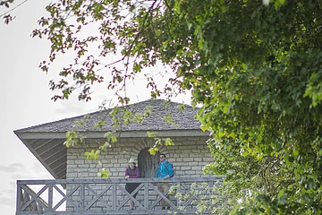 Eine Frau mit Strohhut und ein Mann mit blauer Jacke stehen an dem hölzernen Geländer oben auf dem Limesturm. Hinter ihnen ist eine hölzerne Tür mit der man ins innere des Turms gelangt. Der Turm hat weiter unten noch ein kleines Fenster und links sind einige Steinstufen. Rechts wird der Turm von grünen Blättern der Bäume eingerahmt.