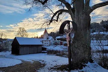Winterweg mit Baum und einem Kreuz. Im Hintergrund Häuser und eine Kirche mit Schnee bedeckt.