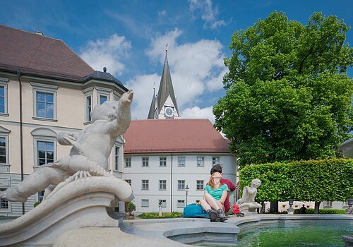 Paar sitzt am Rand eines Brunnens mit Steinfiguren vor historischen Gebäuden und einer Kirche bei blauem Himmel.