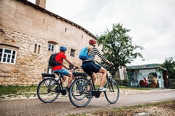 Zwei Radfahrer mit Helmen und Rucksäcken fahren an einer alten Steinmauer und einem Baum vorbei.