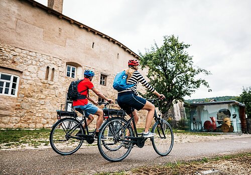 Zwei Radfahrer mit Helmen und Rucksäcken fahren an einer alten Steinmauer und einem Baum vorbei.