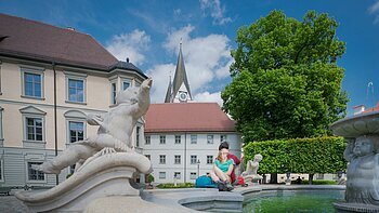 Paar sitzt am Rand eines Brunnens mit Steinfiguren vor historischen Gebäuden und einer Kirche bei blauem Himmel.