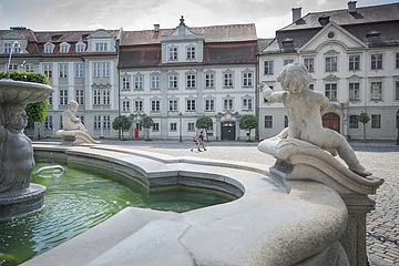 Brunnen mit Puttenfiguren im Vordergrund und historische Gebäude mit roten Dächern im Hintergrund, zwei Personen gehen.