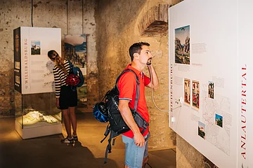 Mann mit rotem Hemd hört an Museumstafel mit Audioguide, Frau liest Ausstellungstext im Hintergrund.