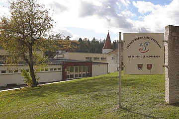 Schild mit Aufschrift „Jugendübernachtungshaus Alte Schule Morsbach“ vor einem Gebäude und Baum auf einer Wiese