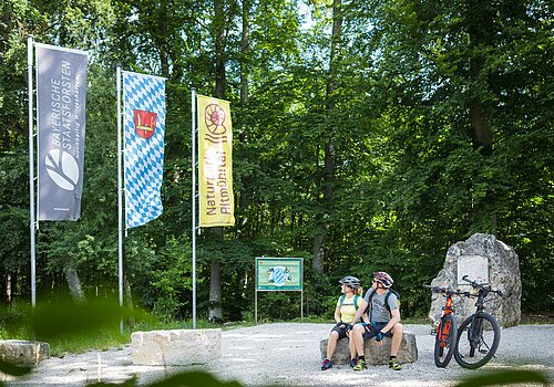 Zwei Radfahrer mit Helm sitzen auf Steinbank im Wald neben zwei Mountainbikes und drei Fahnen.