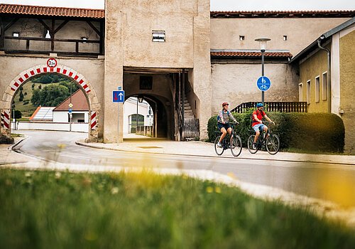 Zwei Radfahrer fahren an einem historischen Stadttor mit rotem Dach und Durchfahrt vorbei.