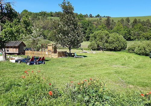 Grüne Wiese mit Bäumen, Blumen, Spielplatz, zwei Kettcars und einem weißen Wohnmobil unter blauem Himmel.